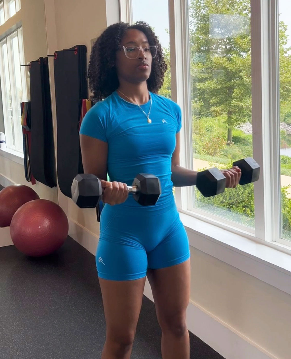 The coach holds a pair of dumbbells at her sides in a bright gym.